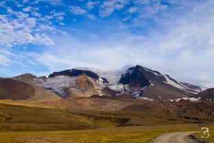 A view of jagged, snow-capped mountains, rolling green-brown terrain, and a glimpse of a road in East Iceland on a blue-sky day.