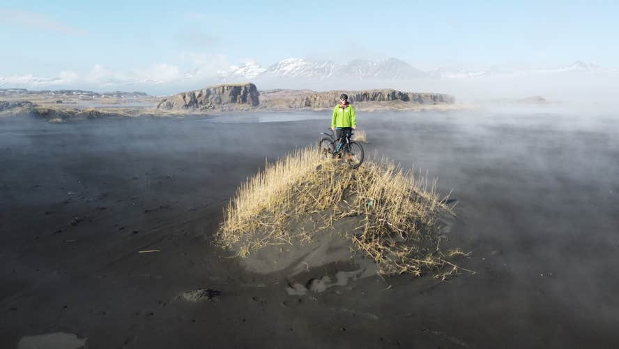 A person stands on a sand mound at the beach, with dramatic hilly scenery behind them.