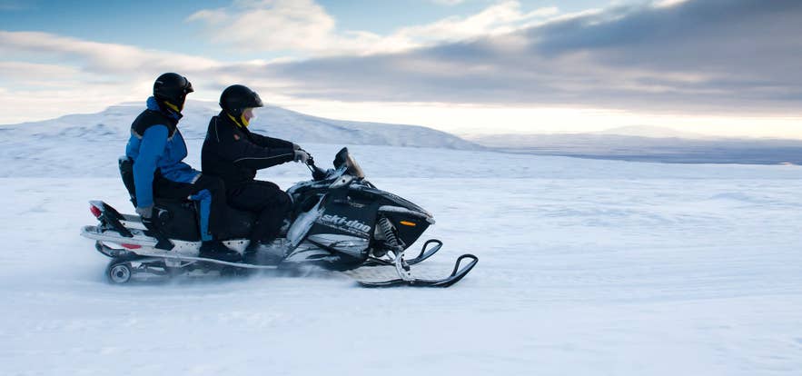 Twee mensen rijden op een sneeuwscooter over een besneeuwd IJslands landschap, met helm en winterkleding tijdens een gletsjertour.