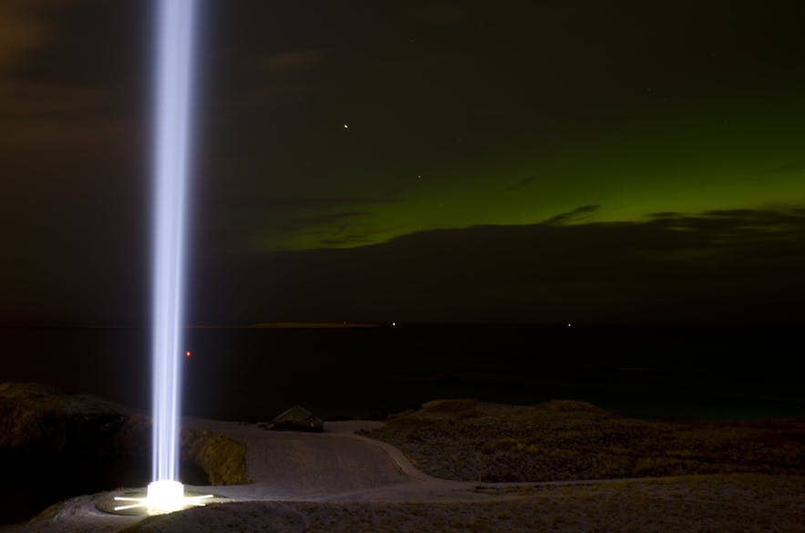 Verticale lichtstraal die omhoog schiet in de nachtelijke hemel op het eiland Videy, met zwakke groene noorderlicht boven de kustlijn.