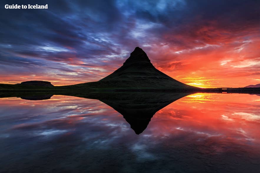 เงาของภูเขาคิร์กจูเฟลล์ (Kirkjufell) สะท้อนบนผืนน้ำที่สงบตัดกับแสงอาทิตย์ยามเย็นใต้เมฆสีแดงและน้ำเงิน
