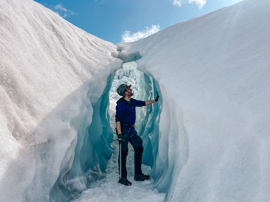 Traveler wearing a helmet and crampons exploring an ice tunnel during a glacier hike in Iceland under a bright blue sky.
