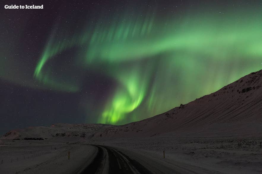 Northern lights over Iceland mountain road with snowy landscape and vivid green aurora dancing in starry night sky