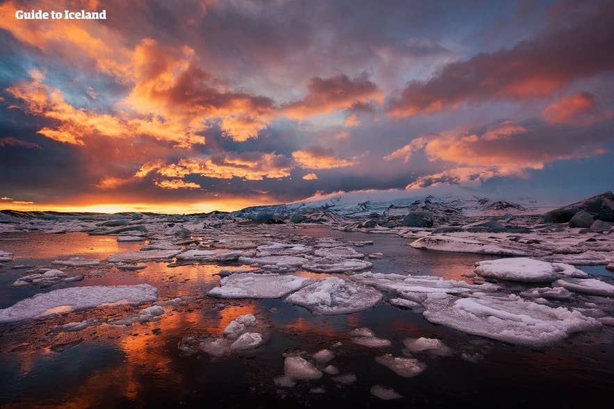 Dramatische zonsondergang boven de Jokulsarlon-gletsjerlagune in IJsland, met oranje wolken die weerspiegelen op het ijzige water vol drijvende, met sneeuw bedekte ijsbergen en een gletsjer in de verte.