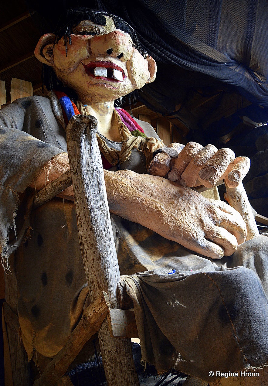 Large sculpture of the Giantess in the Cave, seated with oversized hands and ragged clothes, inside a dimly lit shelter.