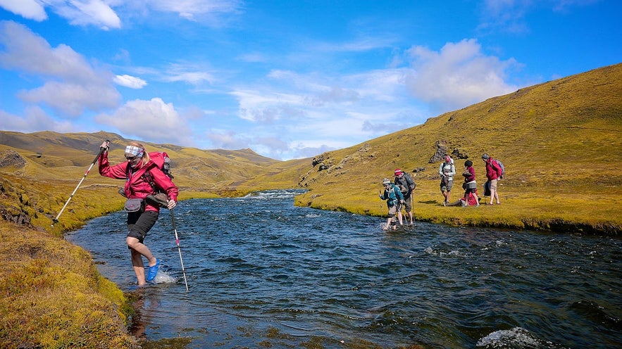 Hikers crossing river in Icelandic Highlands, wearing essential clothes and gear for Iceland tours in summer.