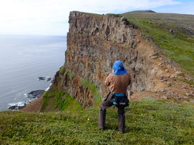 A hiker stands on grassy terrain on top of the green cliffs of Hornstrandir inthe Westfjords.