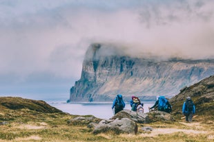 Hikers explore the beautiful Hornstrandir Nature Reserve with calm fjord waters in front and sheer cliffs rising behind, partially enveloped by cloud cover.