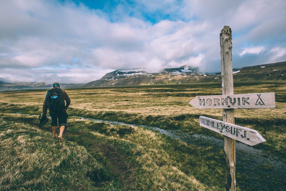 A hiker explore the gorgeous natural landscape of the Hornstrandir Nature Reserve in the Westfjords, with a sign showing directions and snow-speckled mountains rising ahead.