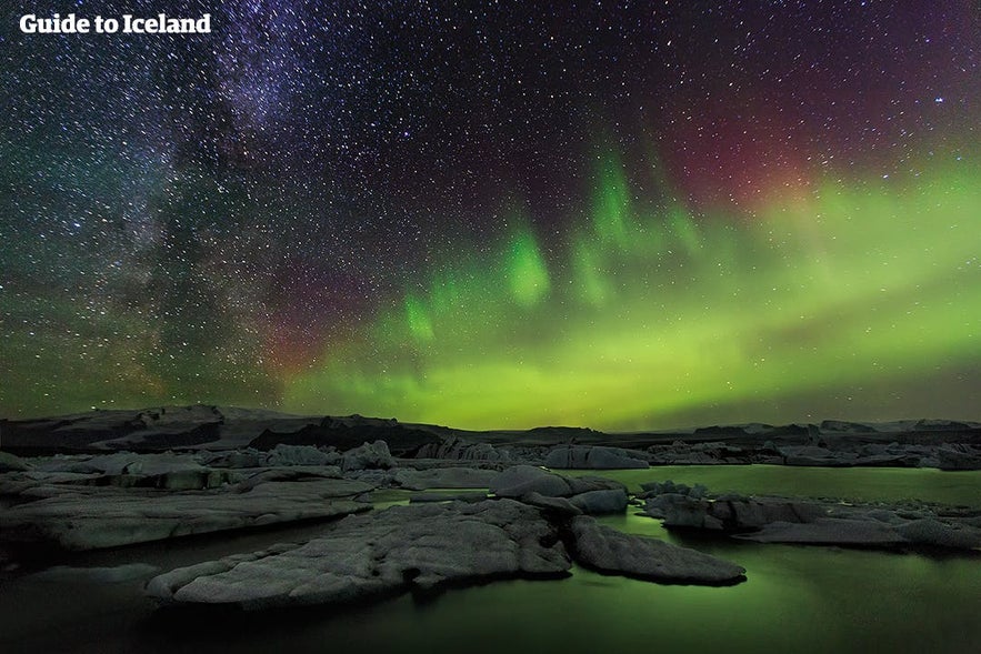 Northern lights in Iceland dancing across the night sky above rocky landscape with reflections on calm water.