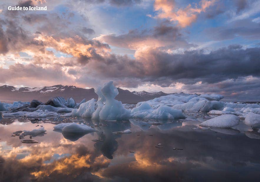 Schitterend uitzicht op drijvende ijsbergen bij de Jokulsarlon-gletsjerlagune in IJsland tijdens zonsondergang, met kleurrijke wolken die weerspiegelen op het stille water en besneeuwde bergen op de achtergrond.