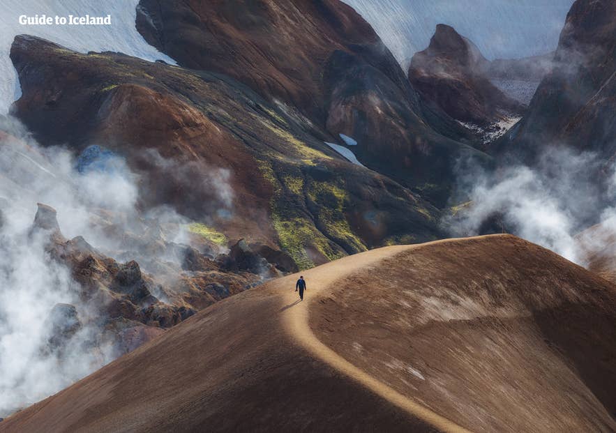 A hiker walks along a narrow ridge in Landmannalaugar, surrounded by colorful rhyolite mountains and rising geothermal steam in Iceland’s highlands.