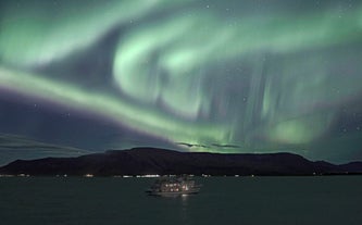 Ein Boot, das auf dem Meer segelt, mit einem Berg im Hintergrund und darüber tanzenden Nordlichtern.
