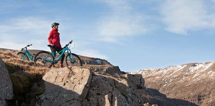 A woman stands beside a mountain bike on top of a cliff wearing a helmet and rainproof red jacket.