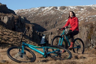 A woman sits on a mountain bike in a grass field with mountains in the background wearing a rainproof red jacket.