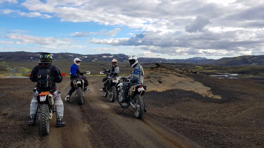 Four riders on adventure motorcycles pause on a Highland gravel track under a big cloudy sky