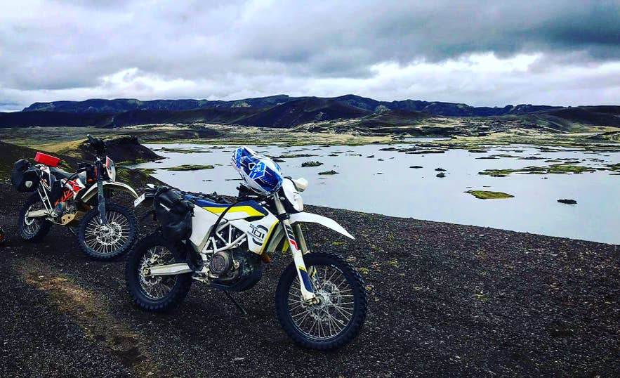 Two adventure motorcycles parked on black gravel beside a shallow Highland lake under grey skies