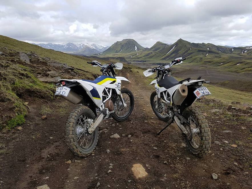 Two white Husqvarna enduro bikes parked on a dirt track facing green mossy Highland mountains