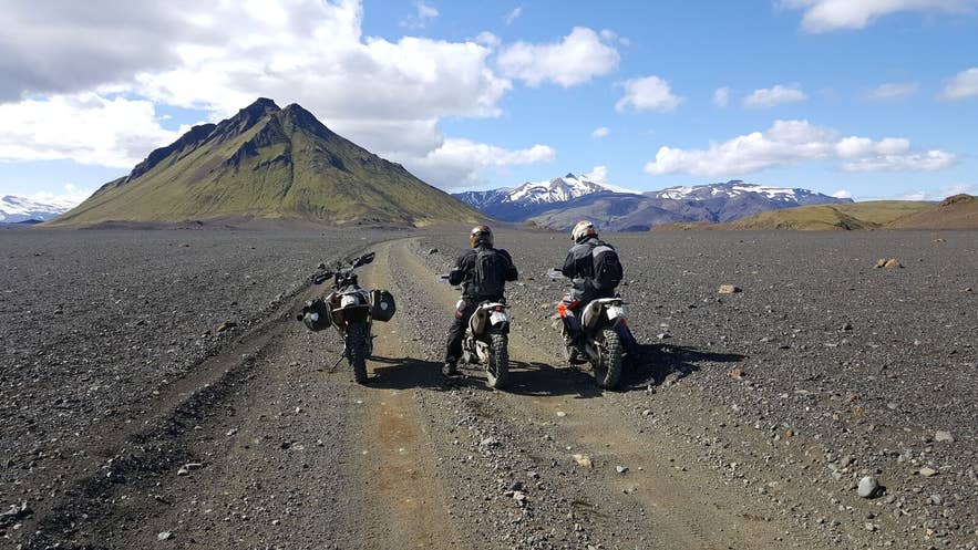 Two riders on enduro bikes on a gravel track crossing a black sand plain toward a green peak