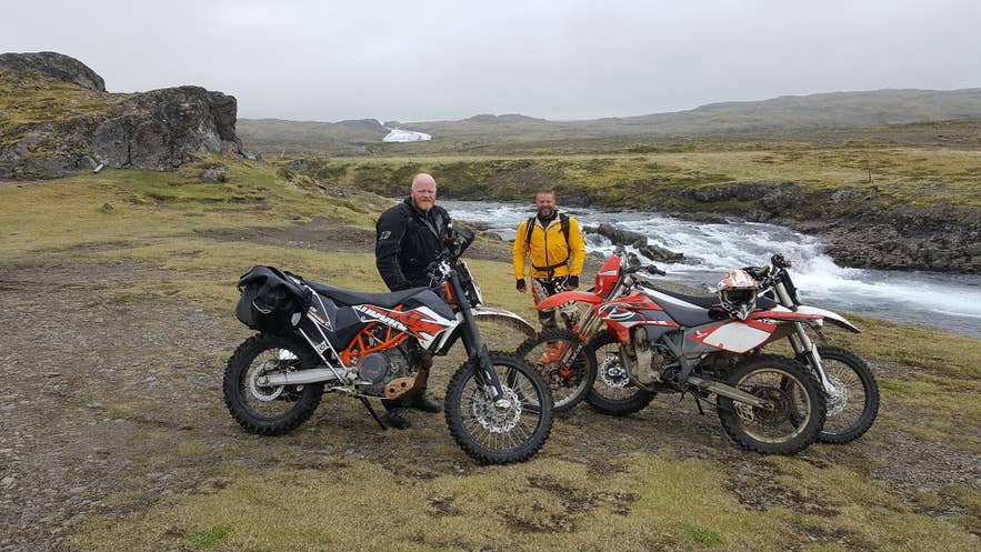 Two riders stand beside enduro motorcycles by a rushing river in green Highland moorland