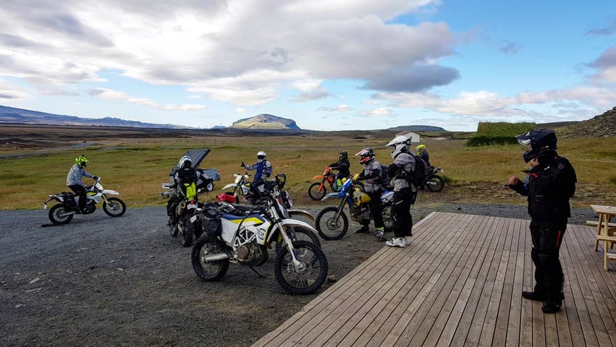 Group of riders with enduro motorcycles gathered by a wooden hut deck on a grassy Highland plain