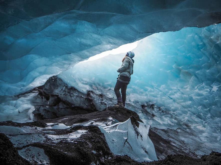 Hiker in a helmet standing on dark rock beneath a blue ice arch inside a glacier. Hiker in a helmet standing on dark rock beneath a blue ice arch inside a glacier.