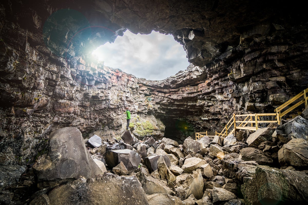 A person explores rocky entrance of Vidgelmir Lava Cave under natural skylight.