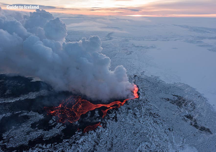 Drohnenaufnahme des Holuhraun-Ausbruchs in Island mit glühenden Lavaströmen und dichten Dampfwolken über einer schneebedeckten Vulkanlandschaft bei Sonnenuntergang.