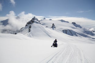 Person kjører snøscooter mot kamera med snødekt fjell i bakgrunn.