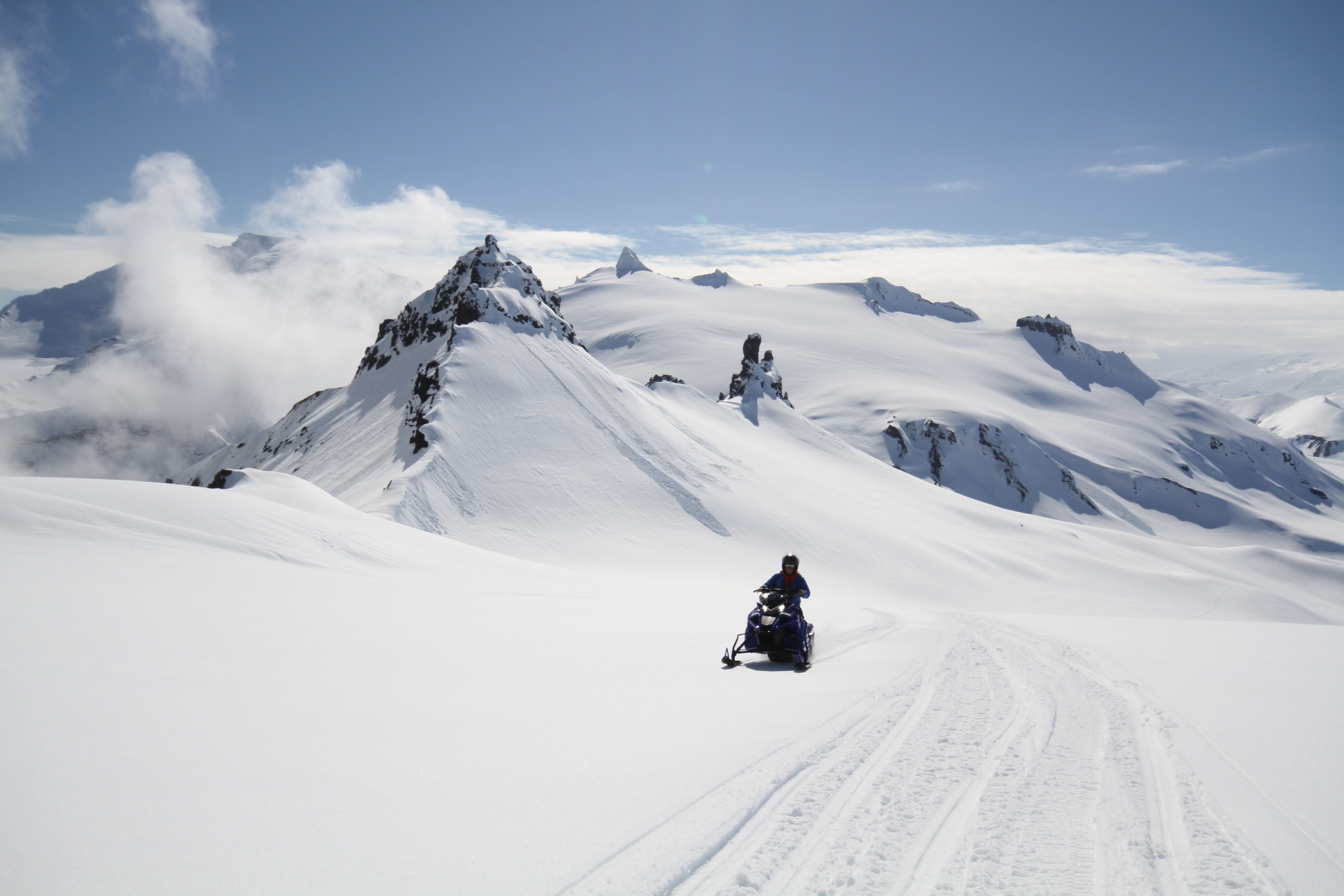 Person kjører snøscooter mot kamera med snødekt fjell i bakgrunn.