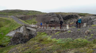 Visitors walk along a rugged footpath and a small bridge that crosses part of the canyon, surrounded by volcanic cliffs and grassy slopes.