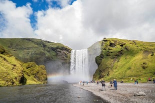 Skogafoss waterfall cascades, with people in front of river bank.