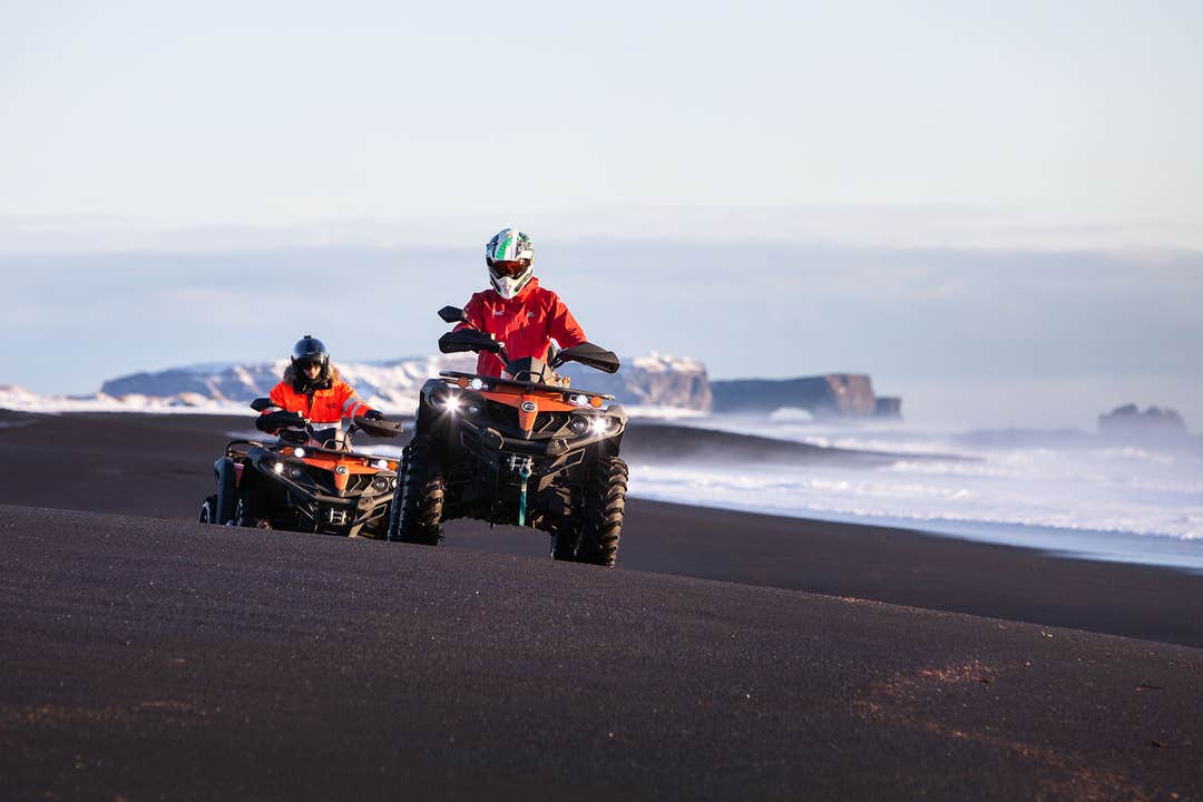 Deux conducteurs de quad, vêtus de vestes rouges, roulent sur une plage de sable noir, avec les vagues de l'océan et des falaises rocheuses en arrière-plan.