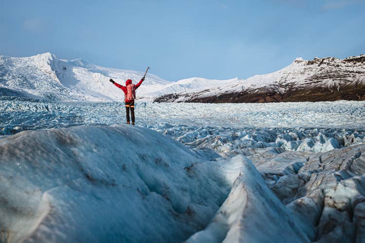 A person on a glacier hiking tour in Iceland raises their hands in the air, holding an ice axe in one hand.