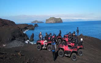 Gruppe på ATV-tur holder pause på en sort sandstrand med havet og klippeformationer foran sig.