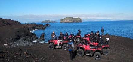 Group ATV tour stopping on a black sand beach with the ocean and rocky outcrops in front.