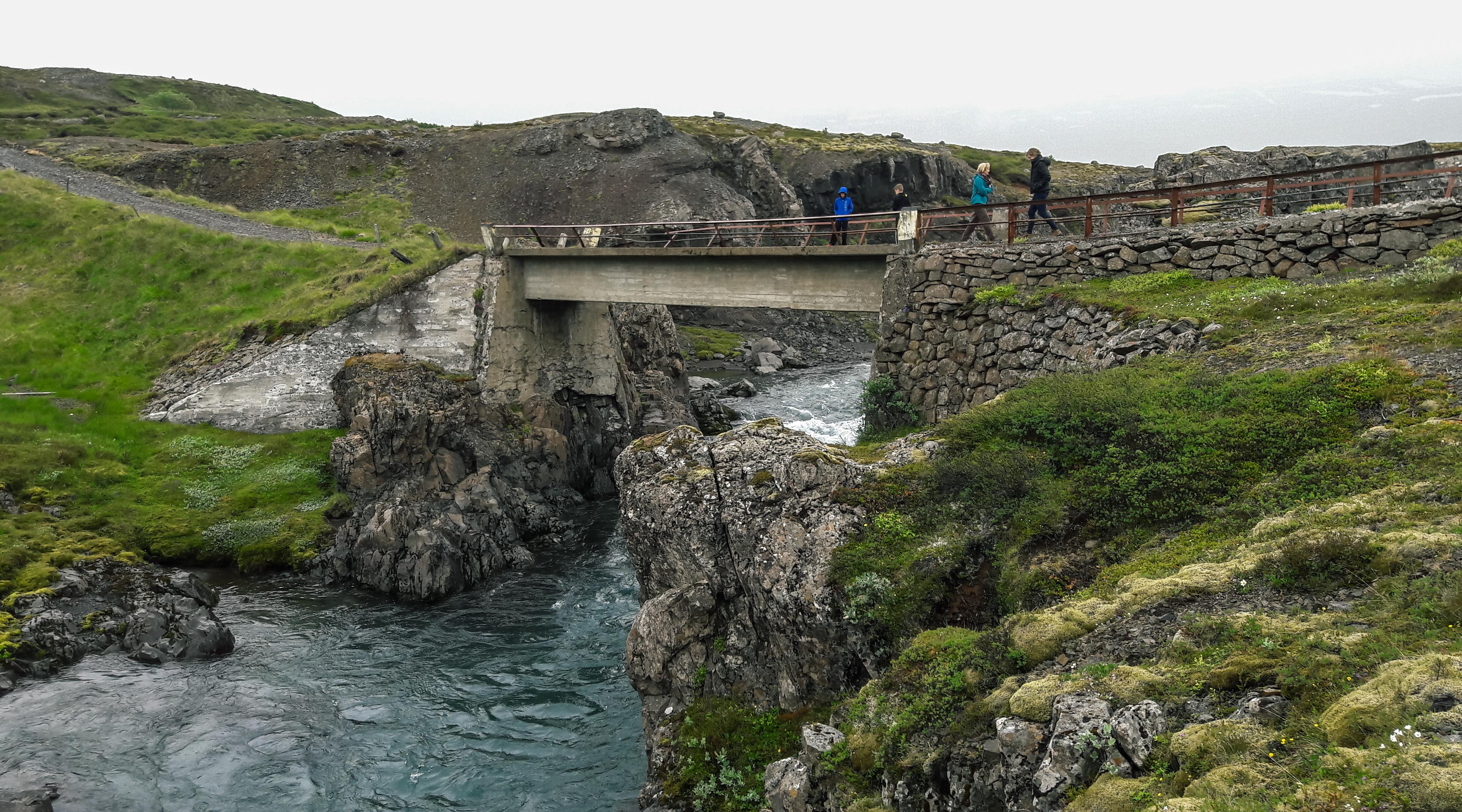 Travelers crossing a scenic bridge spanning a canyon over a river in East Iceland.