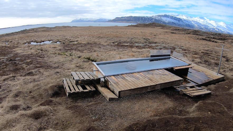 A wooden-decked hot tub on open moorland by a fjord, with snow-capped mountains beyond.