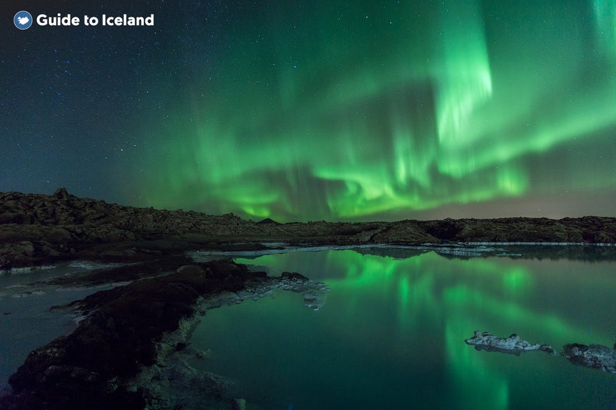 Northern lights over the Blue Lagoon geothermal area in Iceland, with vivid green auroras reflected in the still water under a starry sky.