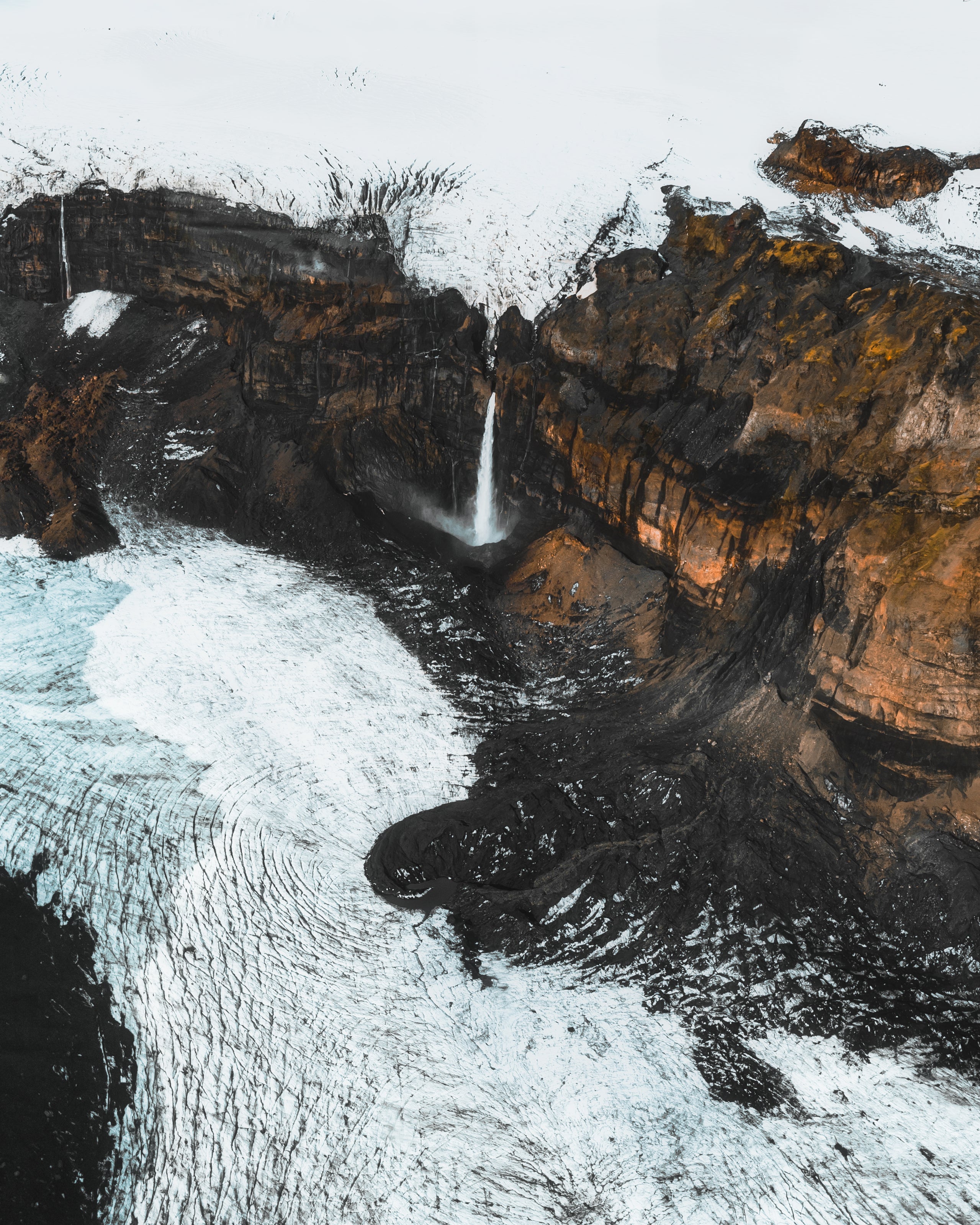A narrow waterfall cascades down a rocky cliff surrounded by snow and ice.