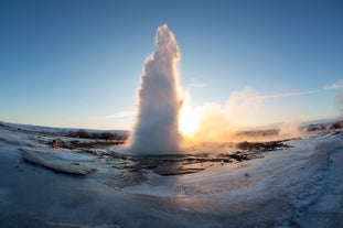 Strokkur geyser erupts with steam and water as the sun rises over the frozen geothermal field in Iceland.