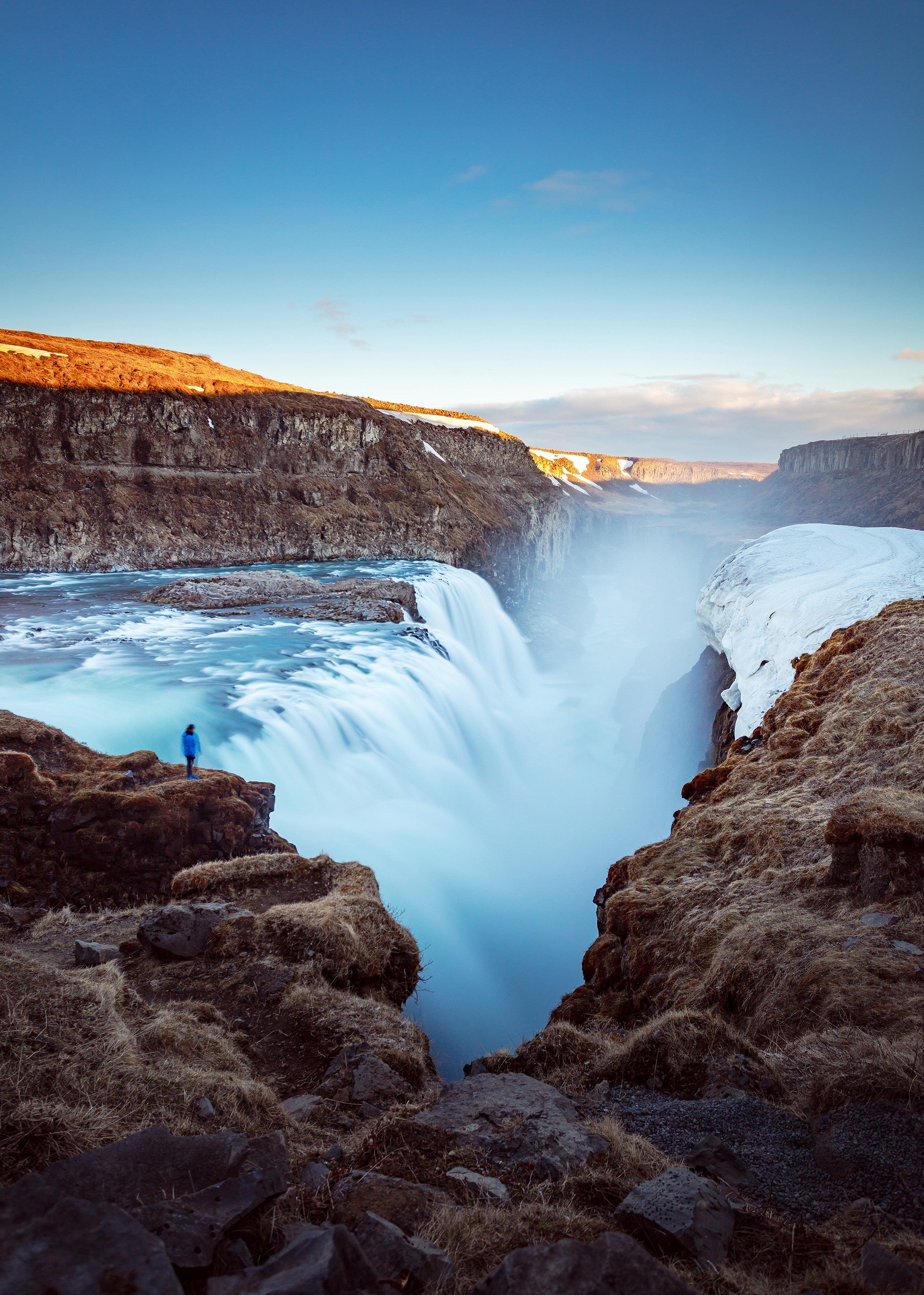 A person stands near Gullfoss waterfall as icy water plunges into a deep canyon on a clear winter day.