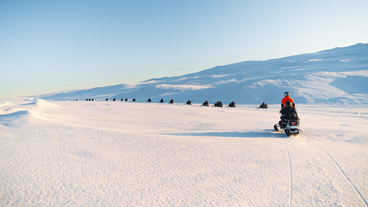 A group of people are seen riding snowmobiles in a line on top of Myrdalsjokull glacier.