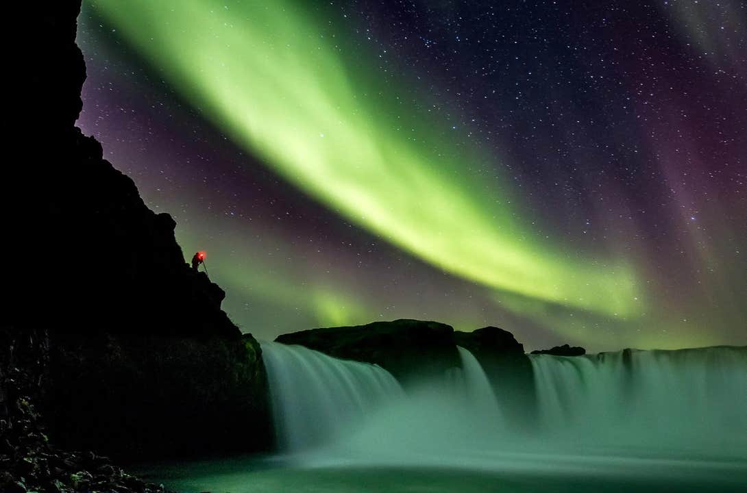 Et nordlysshow danser over et vandfald i Nordisland.