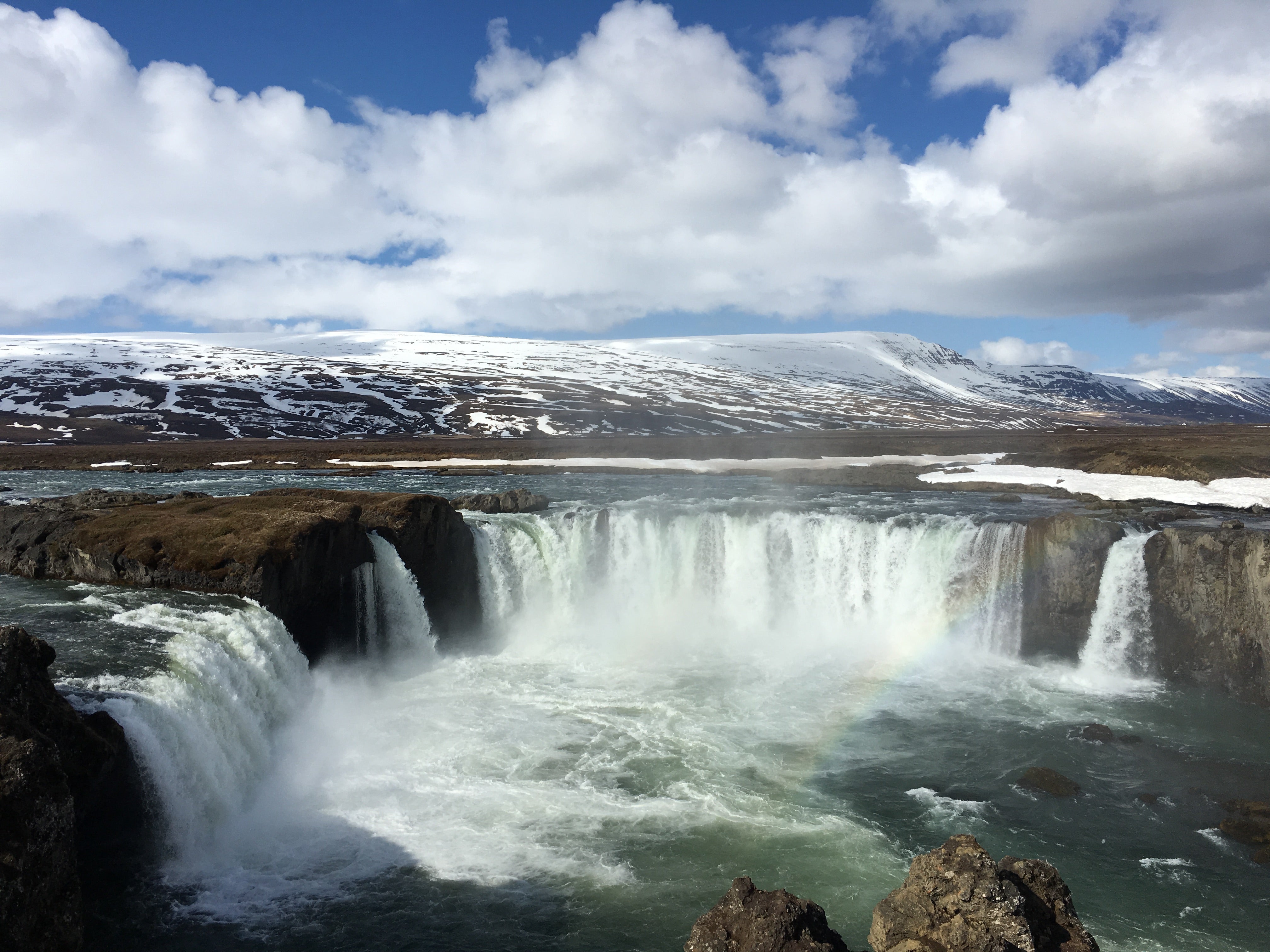 Godafoss Waterfall in spring with snowcapped mountains and a rainbow above the cascade.