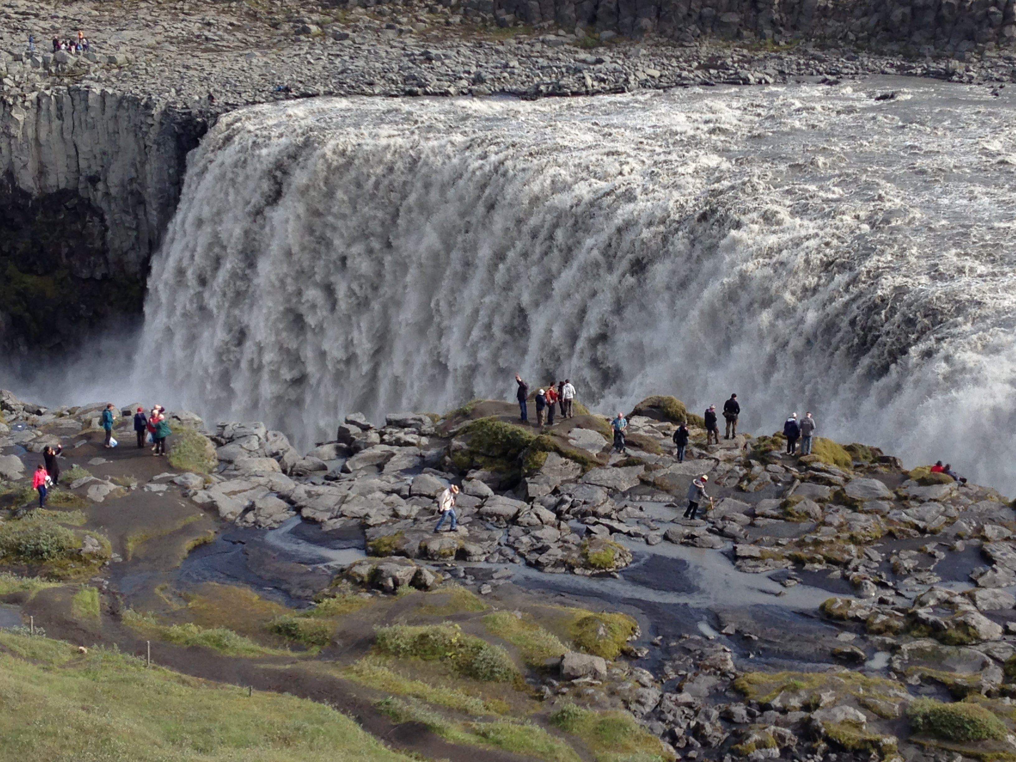 People stand near Dettifoss Waterfall in North Iceland, watching the powerful cascade drop into the canyon.