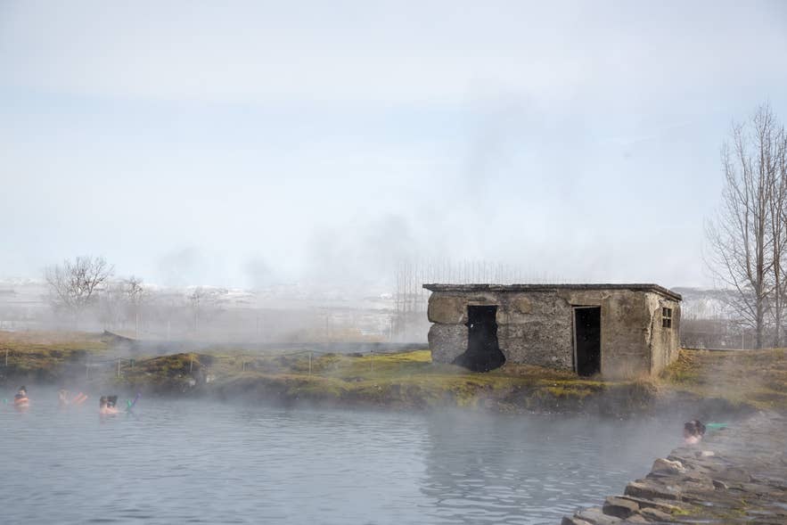 Bathers in the steaming Secret Lagoon by the old stone building, with leafless trees and snowy hills behind