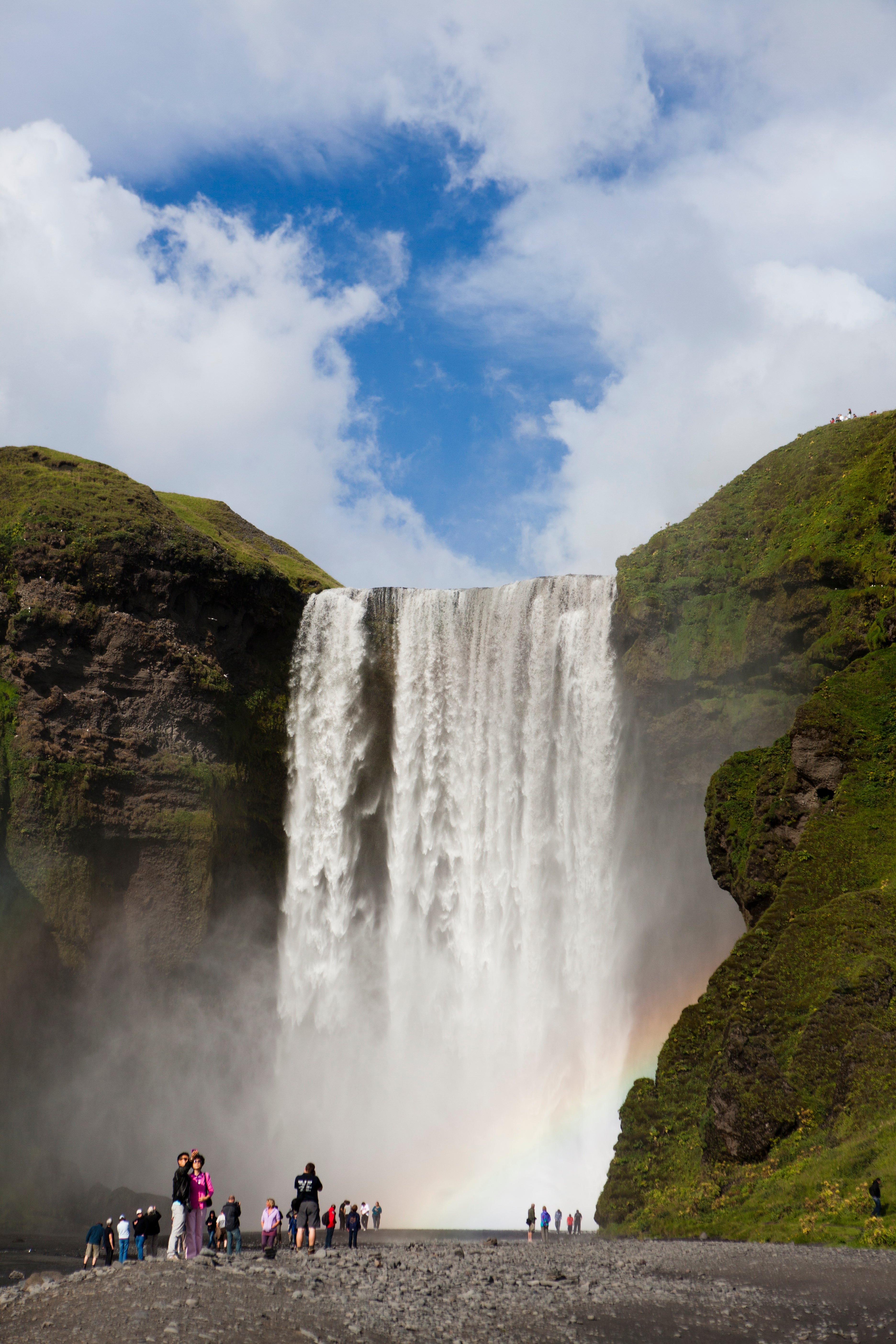 A waterfall in Iceland glimmers on a sunny day, while a rainbow shimmers near the bottom.