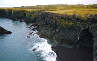 Striking cliffs rise up beside a black sand beach, and the water foams as it reaches the shore.