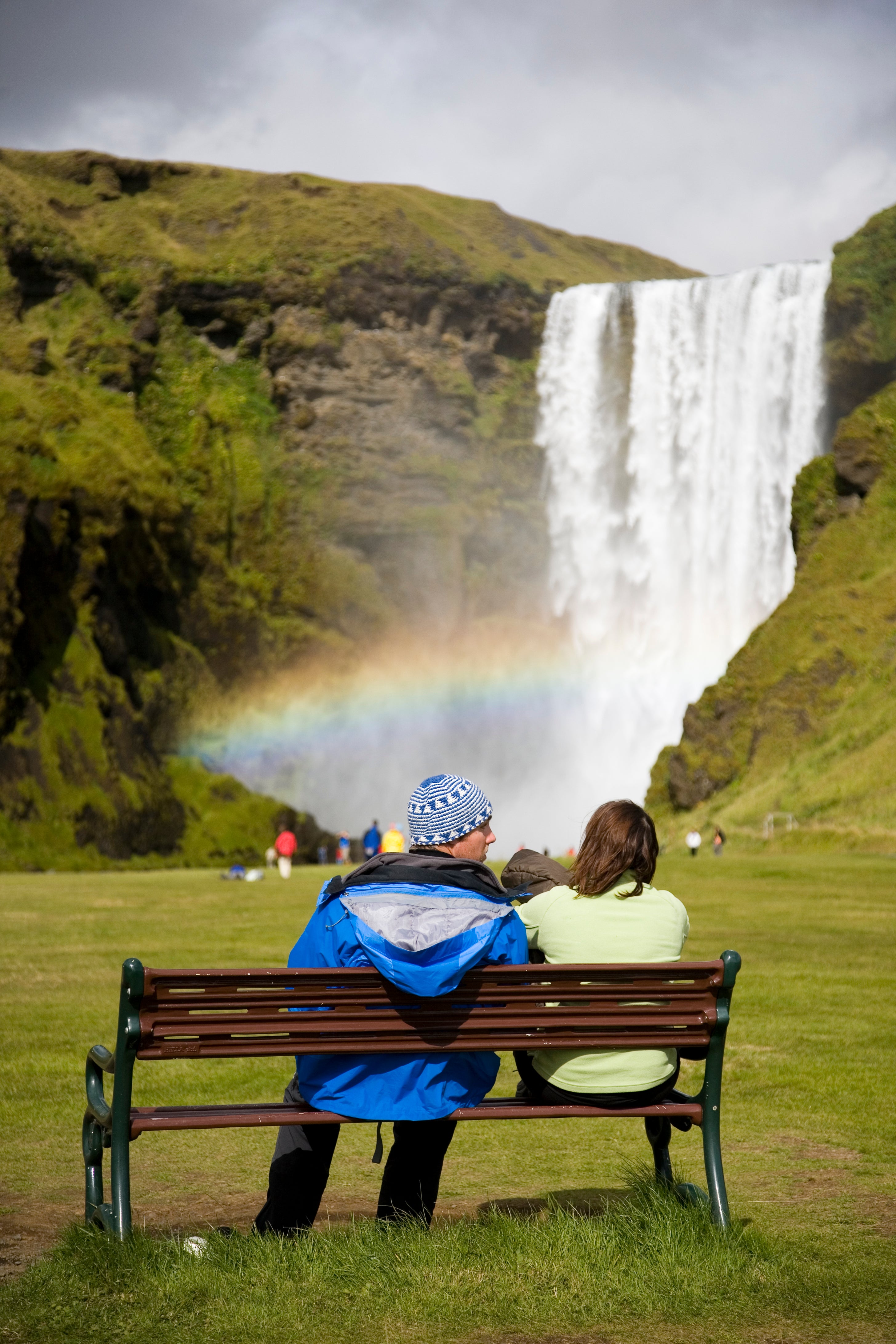 Two people sitting on a bench in front of Skogafoss waterfall which has a rainbow through it.